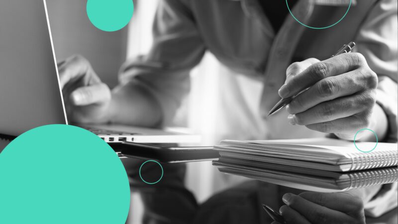 Black and white image of a person's hands typing on a computer and writing in a notepad, with blue circles imposed on top of the image. For the Brightflag blog 